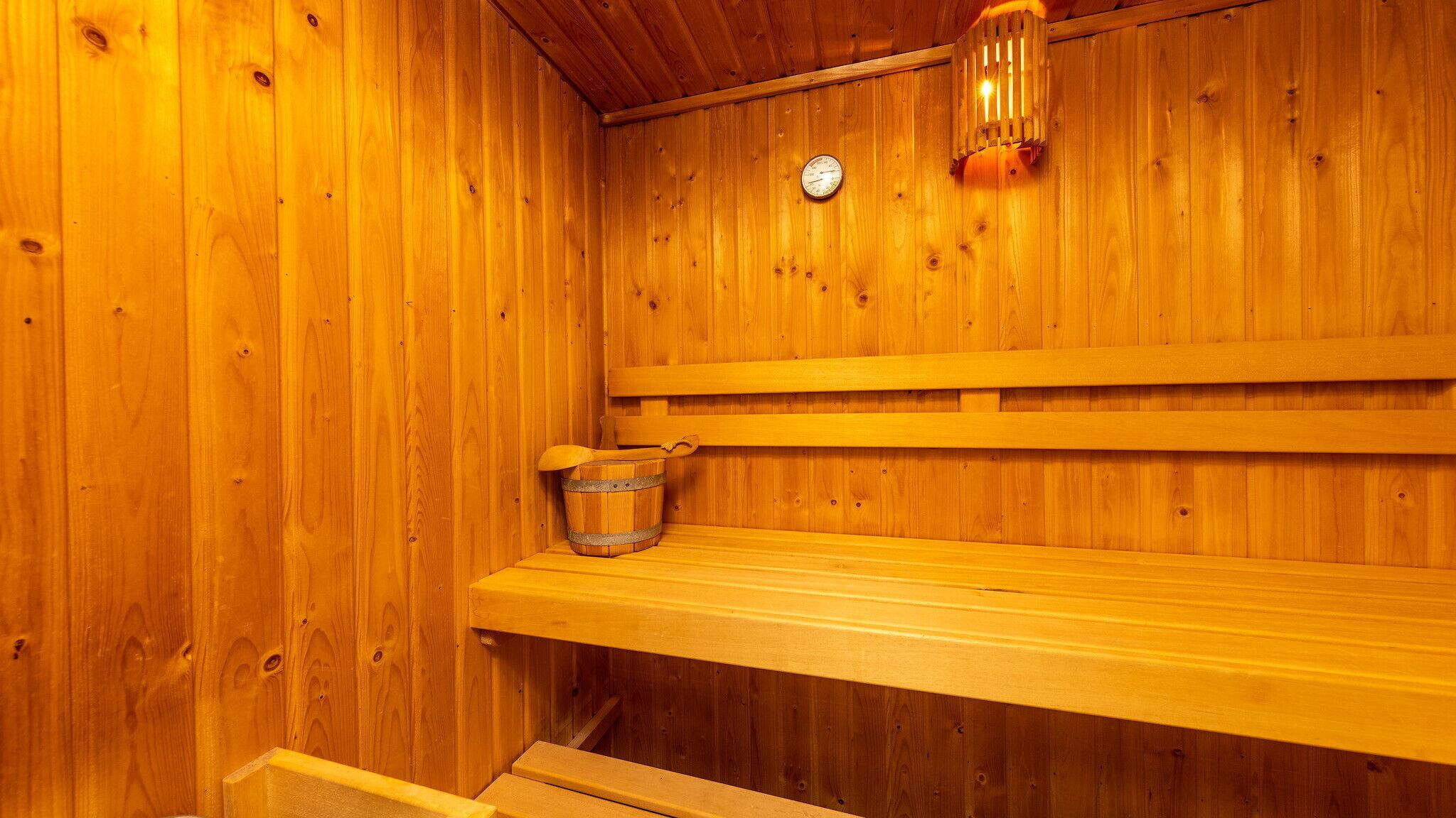 A wooden sauna with a bench, a bucket, and a wall-mounted light fixture.