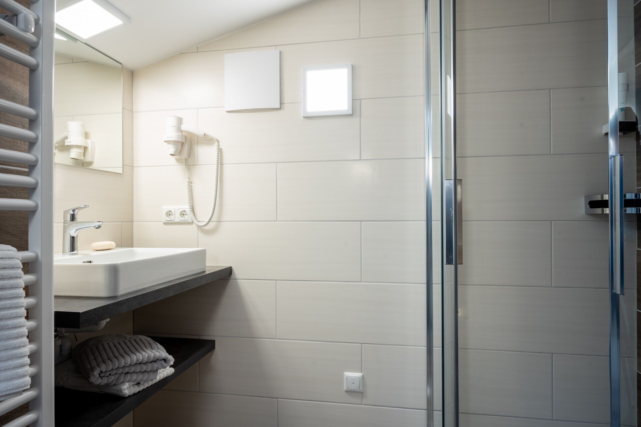 Une salle de bain moderne avec une douche à parois de verre, un lavabo blanc doté d’un porte-savon et un porte-serviettes.
