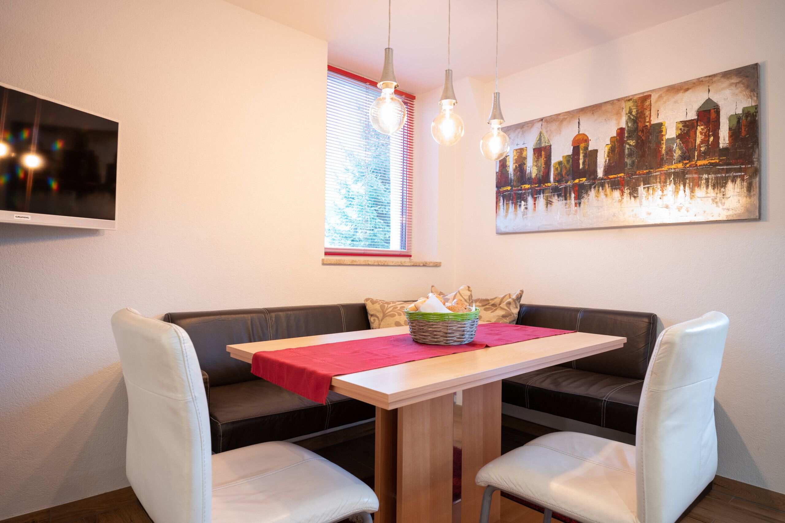 A dining area with a wooden table, white chairs, a red table runner, a basket of bread, and a cityscape painting on the wall.