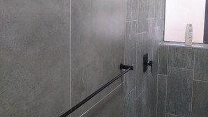 A bathroom with grey tiles, a handrail, a showerhead, and a window with a white blind.