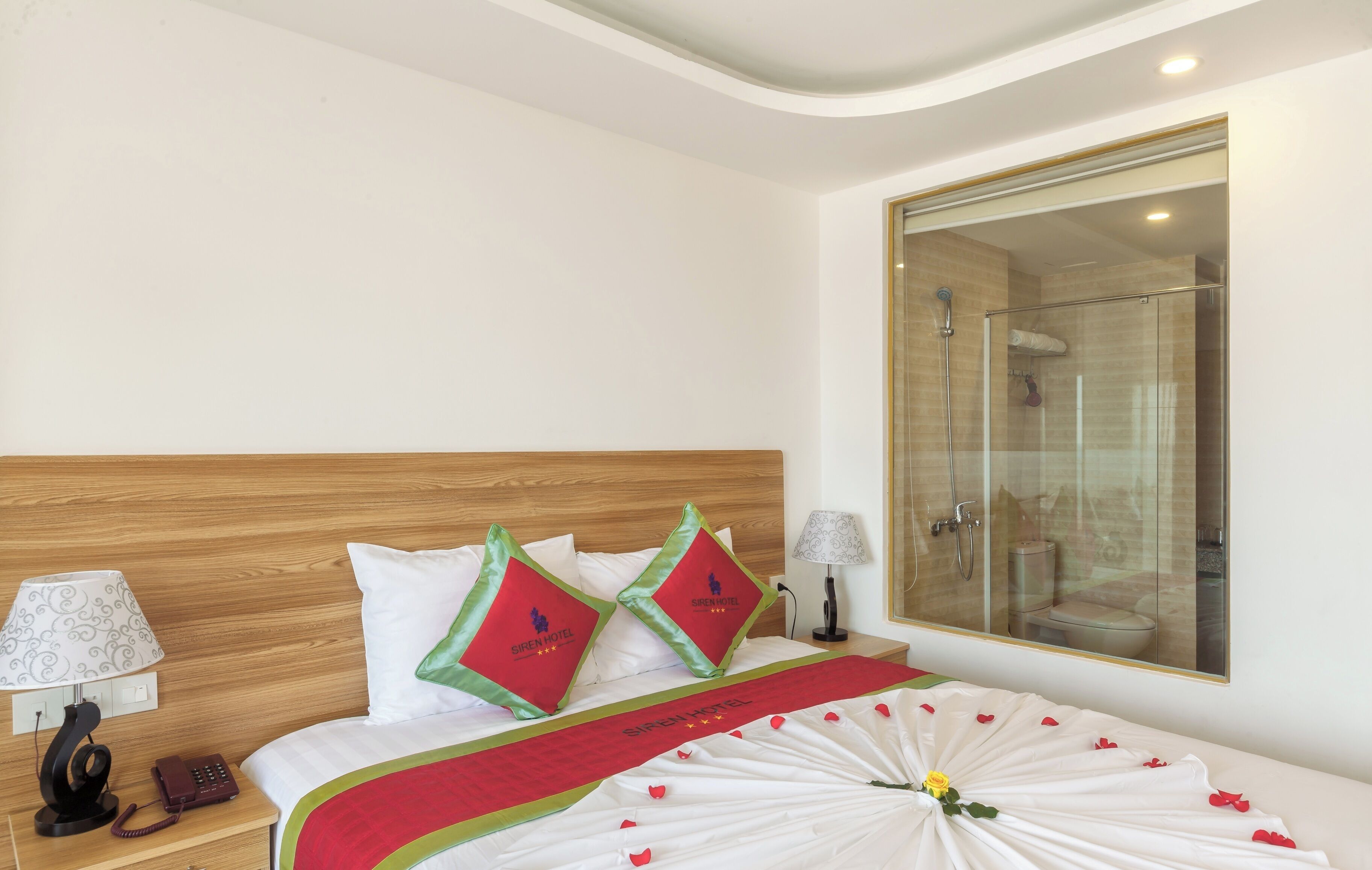 A hotel room with a wooden headboard, two pillows with the hotel logo, a bed with a red and white bedspread, a nightstand with a lamp and phone, and a glass-enclosed shower area.
