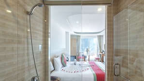 A modern hotel room with a glass shower enclosure, a bed with a white and red bedspread, and a view of the hotel lobby and balcony. - Siren Flower Hotel (Nha Trang)