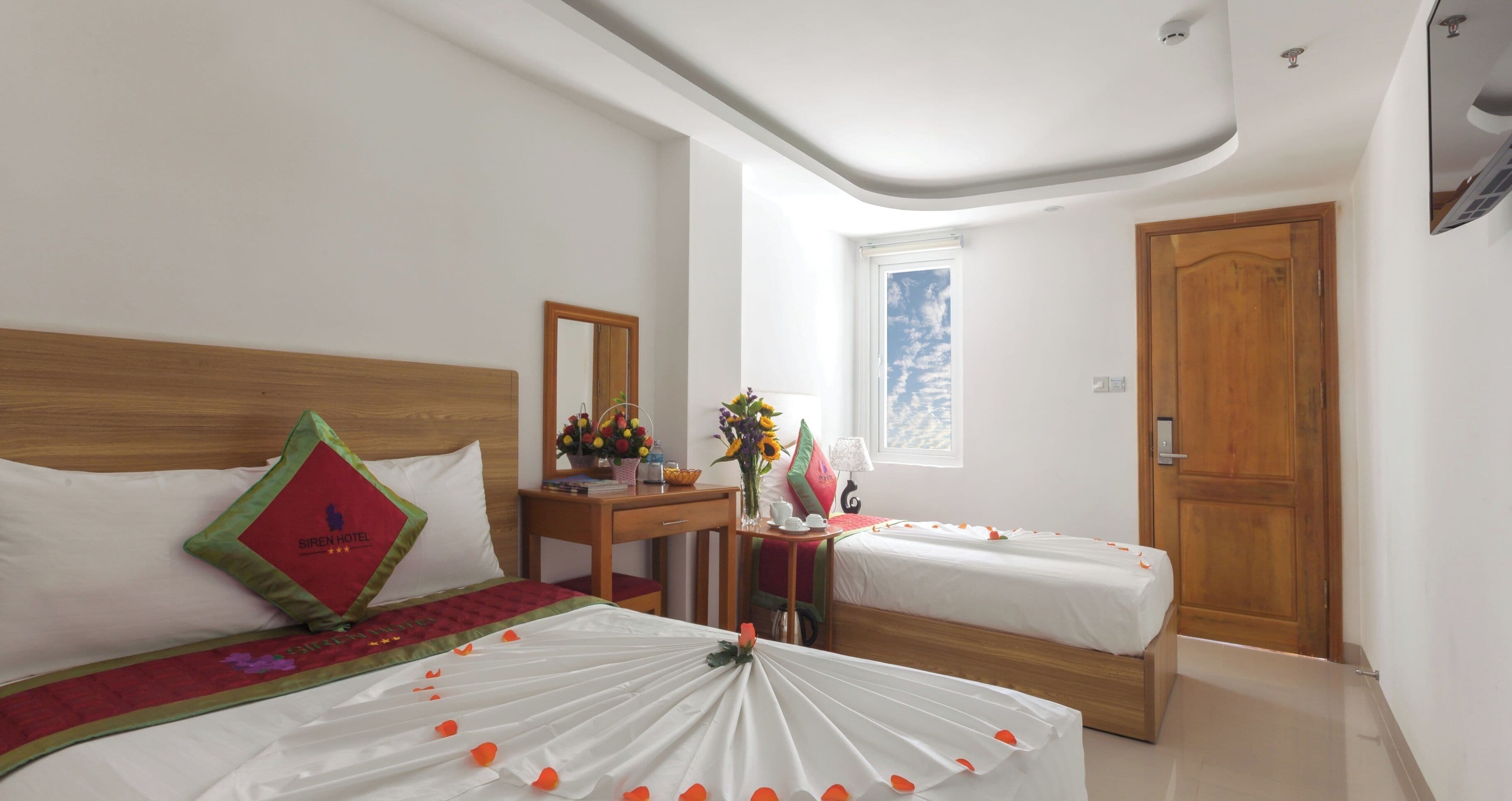 A hotel room with two beds, a wooden headboard, a desk with a mirror, a window with a view, and a wooden door.