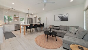 Een moderne keuken met witte kastjes, een eethoek met zwarte stoelen, een grijze bank en een plafondventilator.