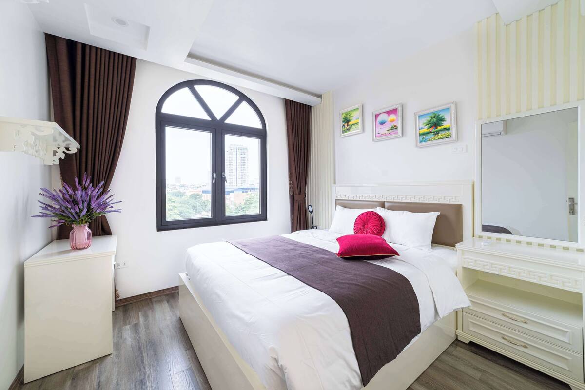 A modern hotel room with a large bed, a bedside table with a vase of flowers, a window with curtains, and a view of the city.
