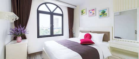 A modern hotel room with a large bed, a bedside table with a vase of flowers, a window with curtains, and a view of the city.