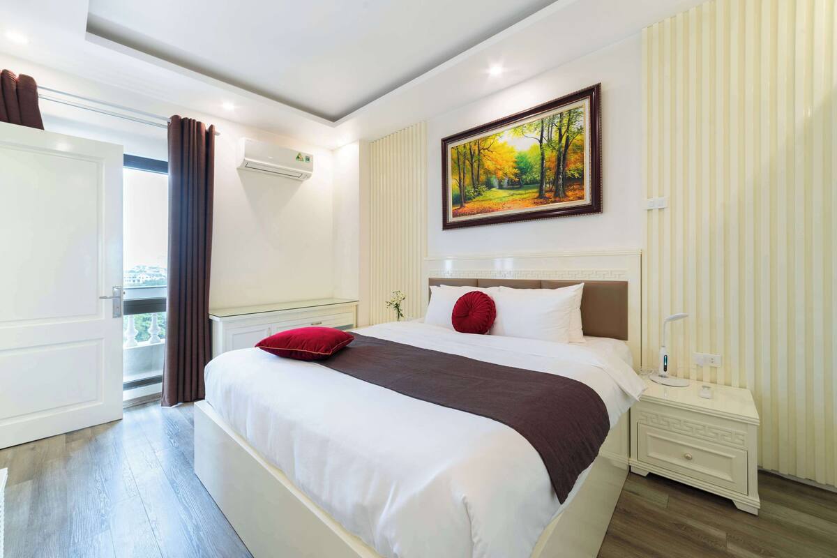 A bedroom with a large bed, a nightstand, a painting on the wall, and a window with curtains.