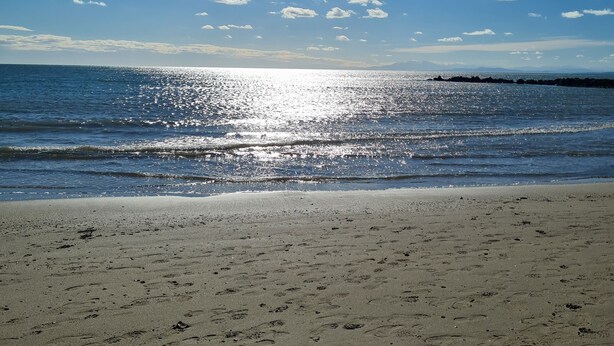 Una playa con huellas en la arena, cielo despejado y el sol reflejándose en el agua.