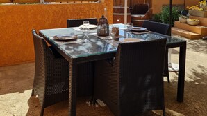 Un patio exterior con una mesa lista para comer, rodeado de sillas de mimbre, bajo una pérgola con flores moradas.