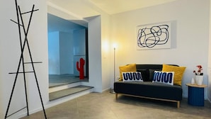 Un salon moderne avec un canapé noir, des coussins jaunes et bleus, un cactus rouge en guise de décoration, et un grand tableau abstrait accroché au mur.