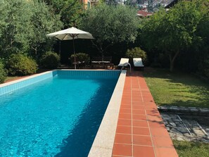 A swimming pool with a tiled edge, surrounded by greenery and a patio area with chairs and an umbrella. - Villa Dell Artista by Elite Villas (Cernobbio)