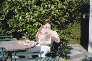 A person reading a newspaper at a round table outdoors with a hedge in the background. - Villa Dell Artista by Elite Villas (Cernobbio)