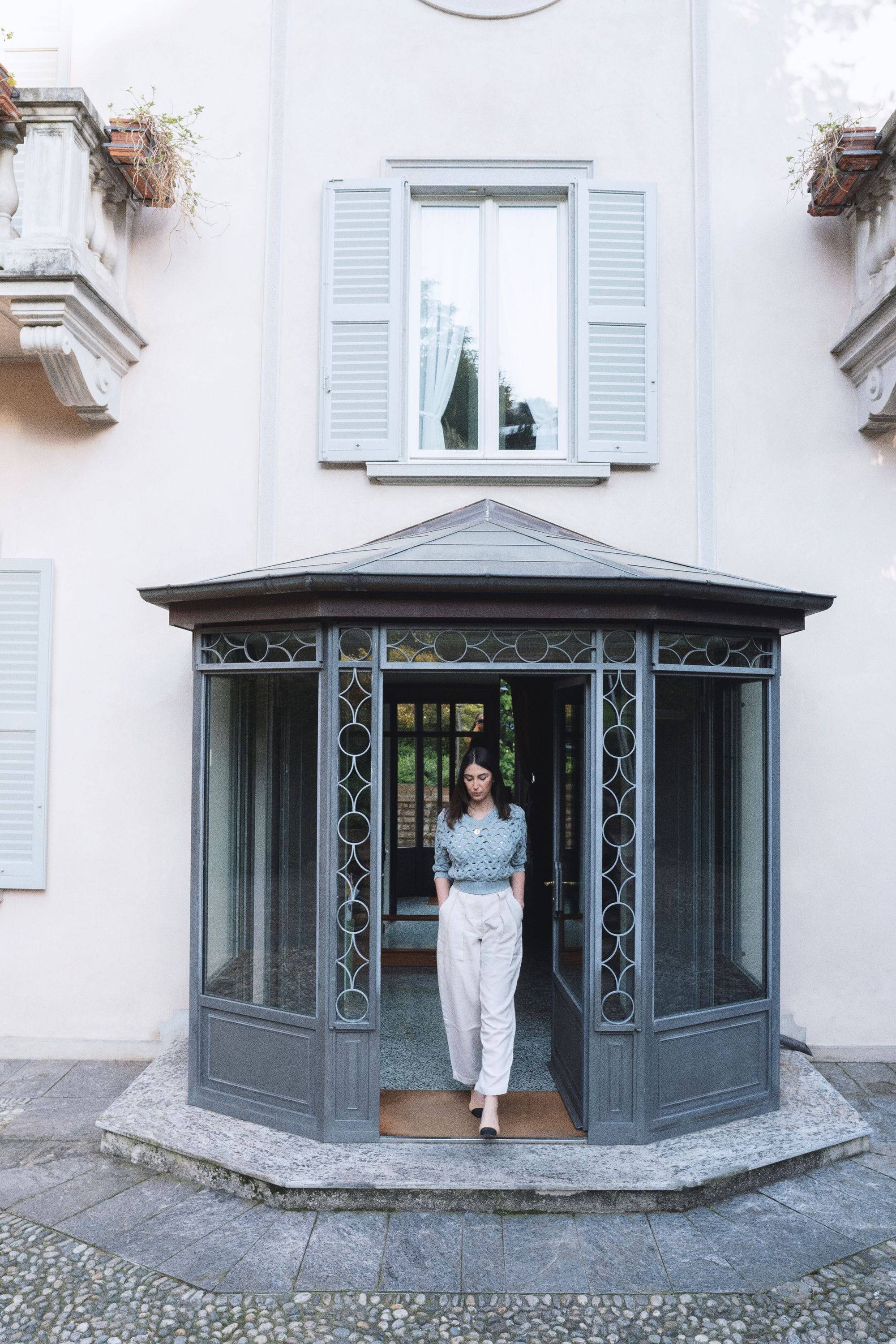 A woman standing in a glass-walled gazebo with a tiled floor, next to a building with white walls and shutters.