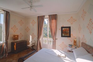 Ein Schlafzimmer mit einem Bett, einem Schreibtisch, einem Stuhl, einer Kommode, einem Deckenventilator und einem Fenster mit Vorhängen.