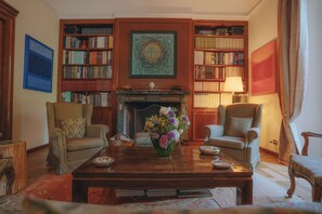 Une pièce avec une cheminée, deux fauteuils, une table basse ornée d’un vase de fleurs et des étagères remplies de livres.