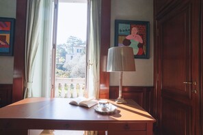Un bureau en bois avec un livre, une tasse de thé et une lampe. Il y a une fenêtre avec des rideaux, une porte et un tableau encadré au mur.