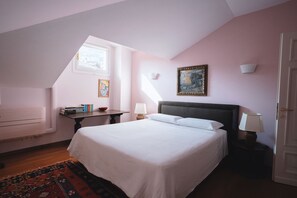 Une chambre avec un grand lit, une petite table avec une lampe, un tableau au mur et une fenêtre laissant entrer la lumière naturelle.