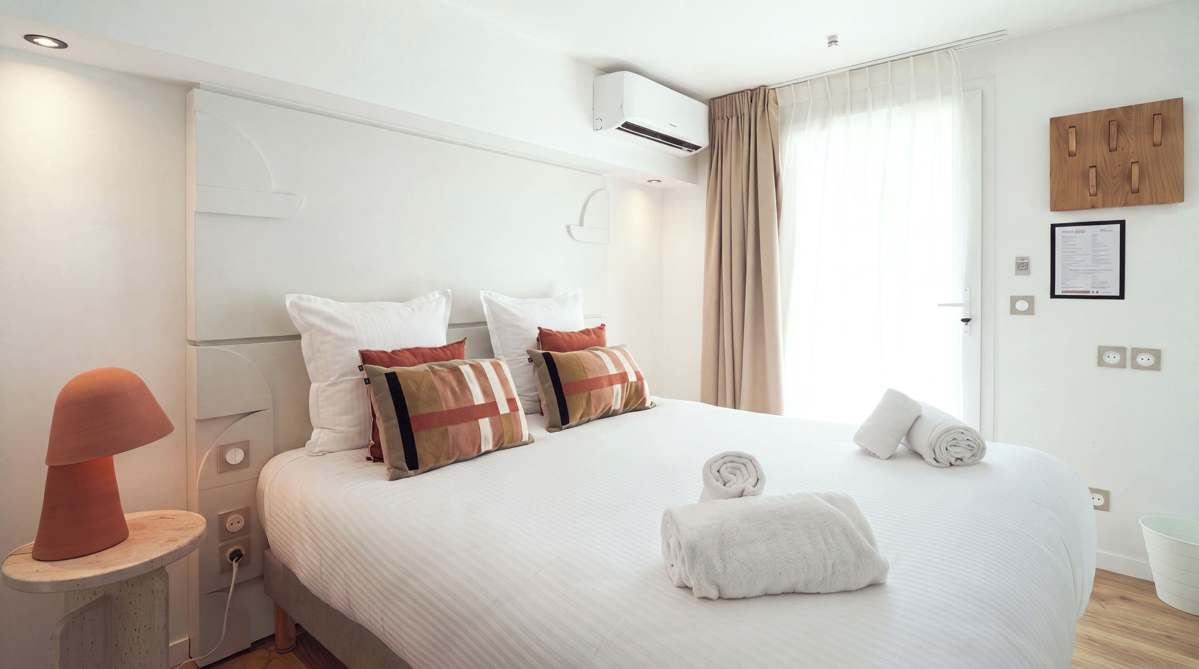 A hotel room with a bed, pillows, a bedside table with a lamp, an air conditioning unit, and a window with curtains.