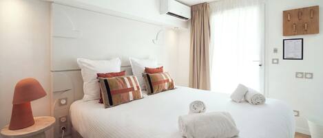 A hotel room with a bed, pillows, a bedside table with a lamp, an air conditioning unit, and a window with curtains.
