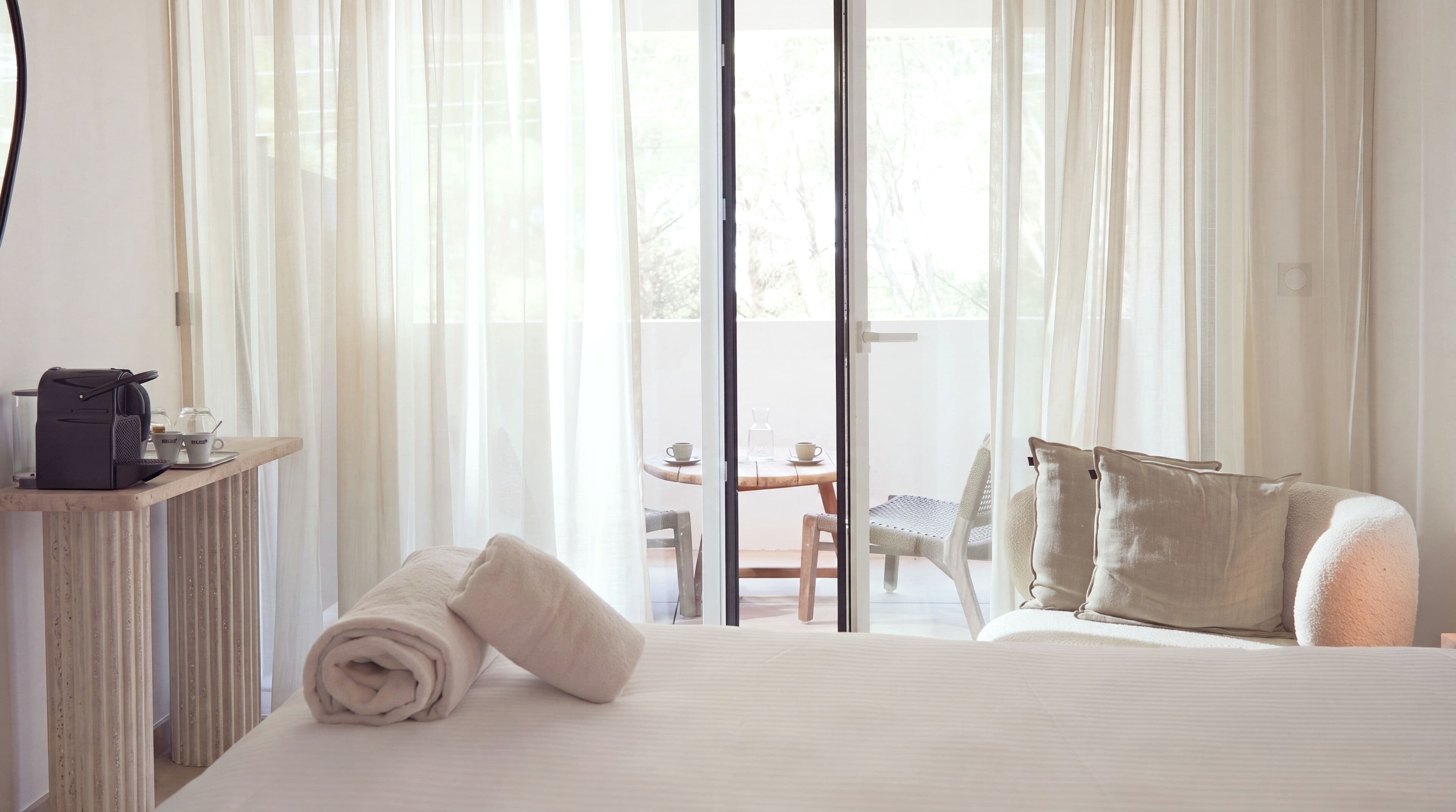 A hotel room with a bed, a desk with a coffee maker, a table with chairs, and a view of the outdoors through large windows.