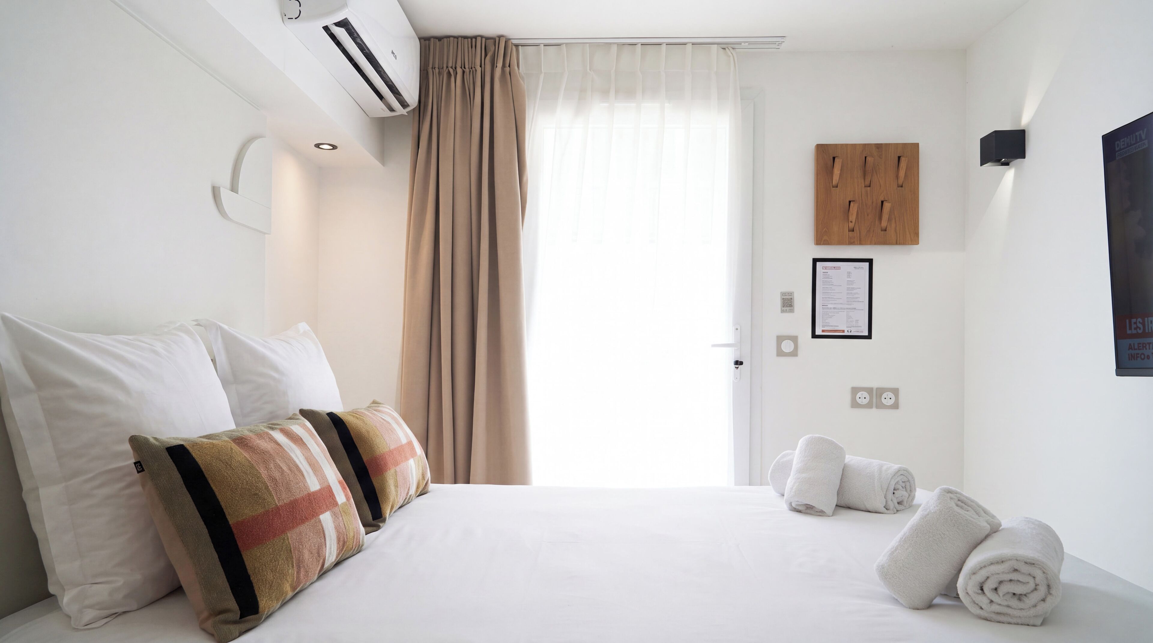 A hotel room with a bed, pillows, a window with curtains, a wall-mounted air conditioner, a wooden panel, a framed notice, and a towel rack.
