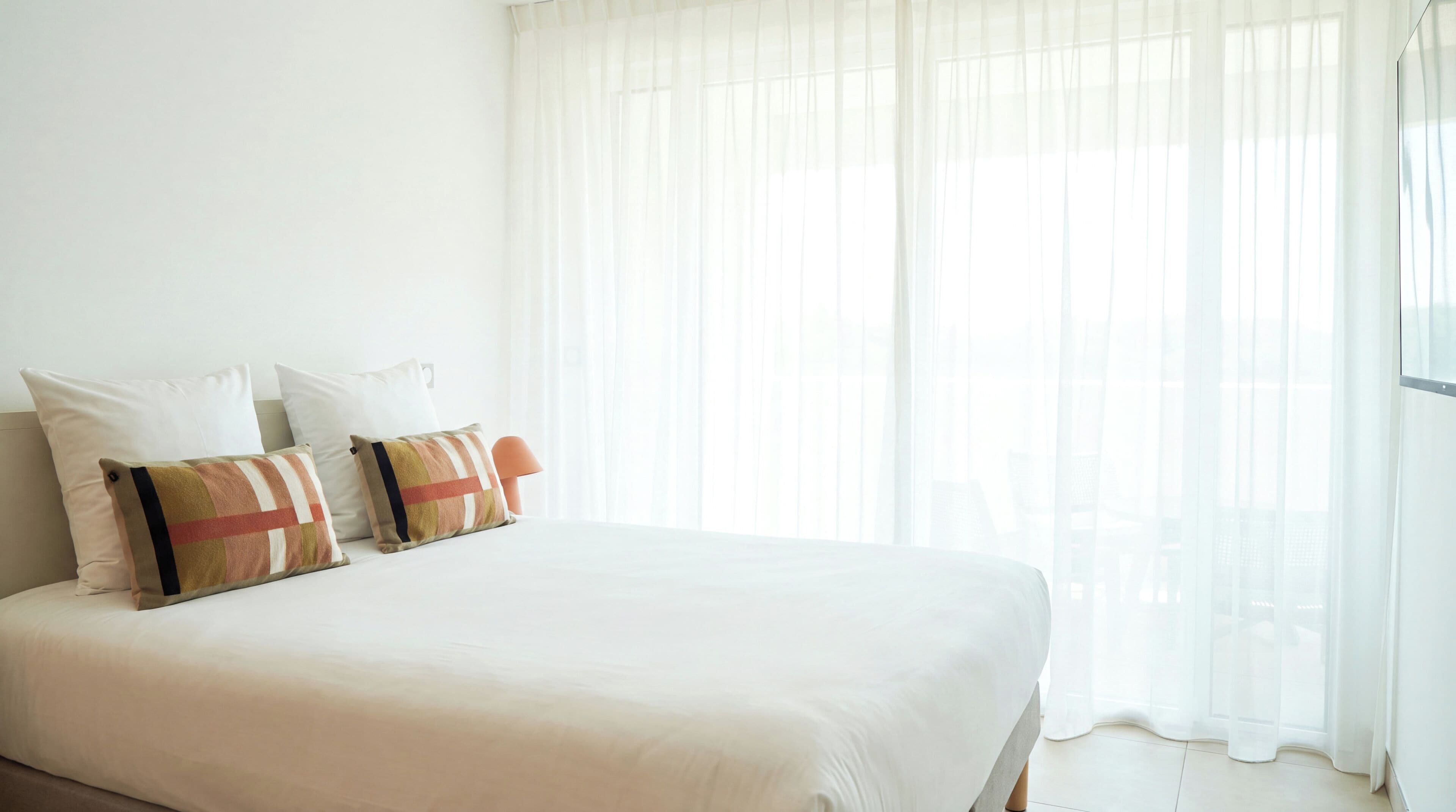 A bed with white bedding and two decorative pillows, a bedside table, and sheer curtains allowing natural light.