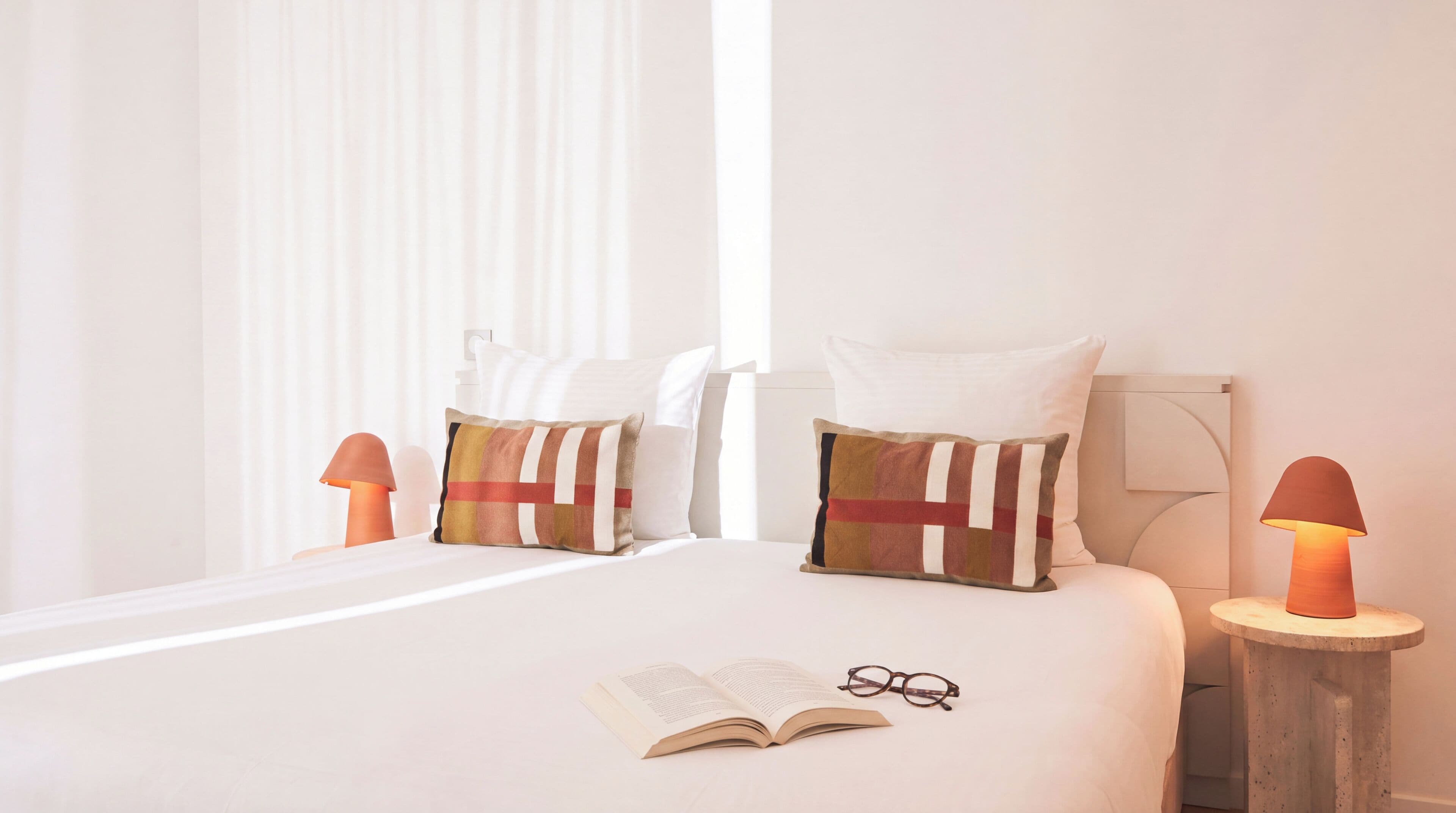 A neatly made bed with a book and glasses on it, two bedside lamps, and a window with sheer curtains.