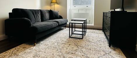 Un salon avec un canapé noir, un tapis à motifs, une petite table, un téléviseur posé sur un meuble et une fenêtre donnant sur un paysage agréable.