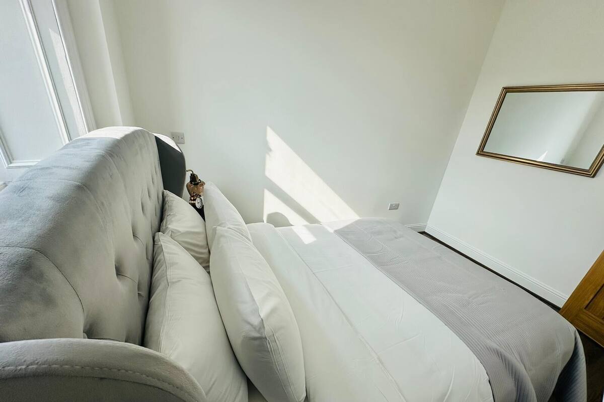 A modern bedroom with a tufted headboard sofa, a bed with a grey headboard, and a large mirror on the wall.