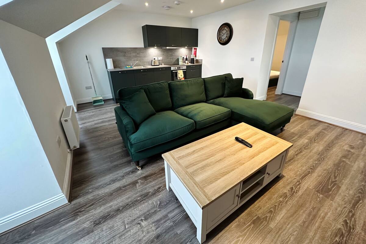 A modern living room with a green sectional sofa, a wooden coffee table, and a kitchen in the background.