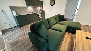 Una sala moderna con un sofá seccional verde, una mesa de centro de madera y una zona de cocina con armarios oscuros y un reloj en la pared.