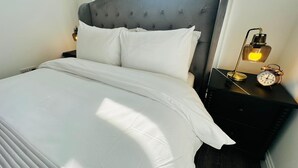 Una cama bien hecha con sábanas blancas, un cabecero capitonado y una mesita de noche con una lámpara y un despertador.