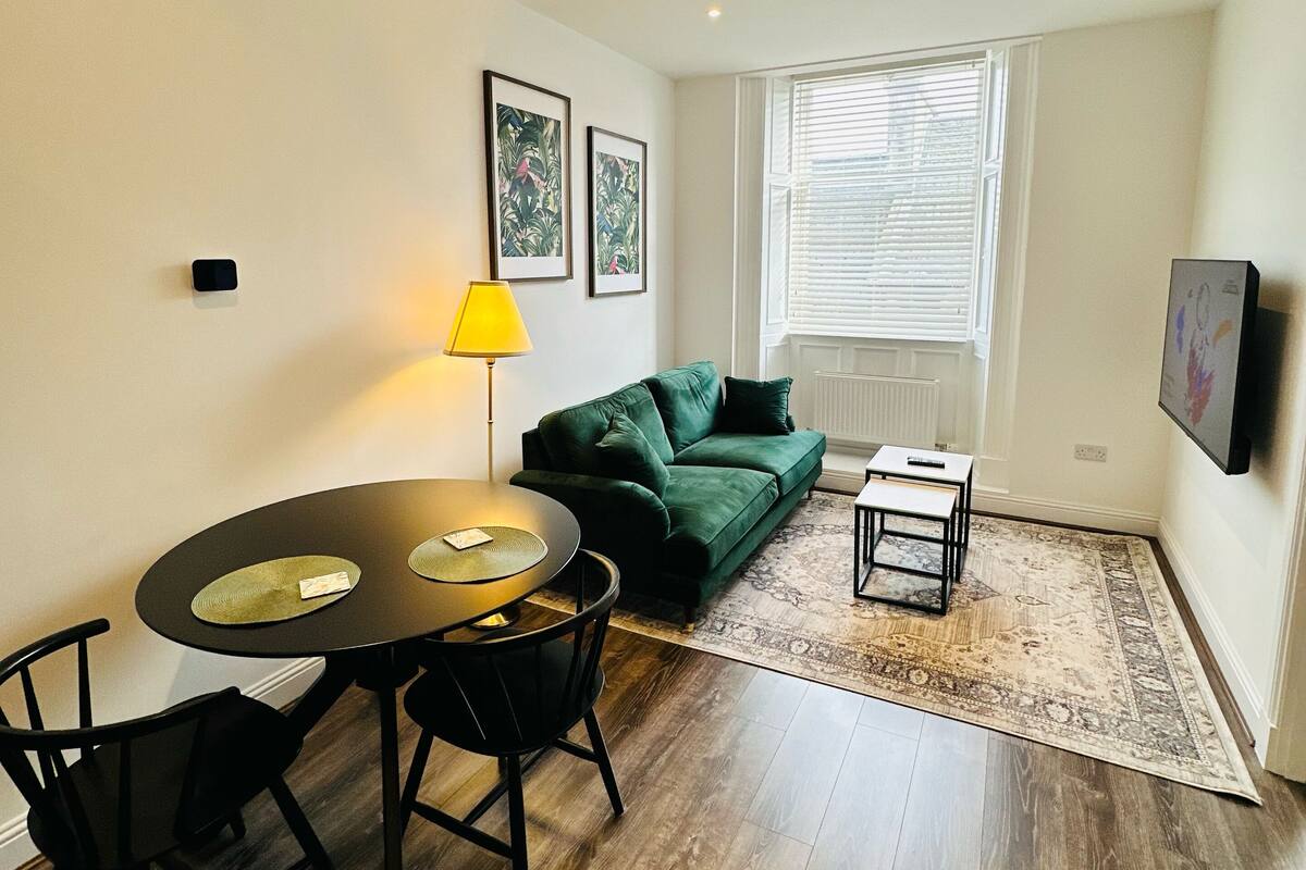 A compact living space with a dining table, chairs, a green sofa, a small side table, and a patterned rug.