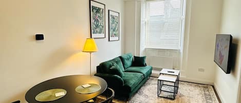 A compact living space with a dining table, chairs, a green sofa, a small side table, and a patterned rug.