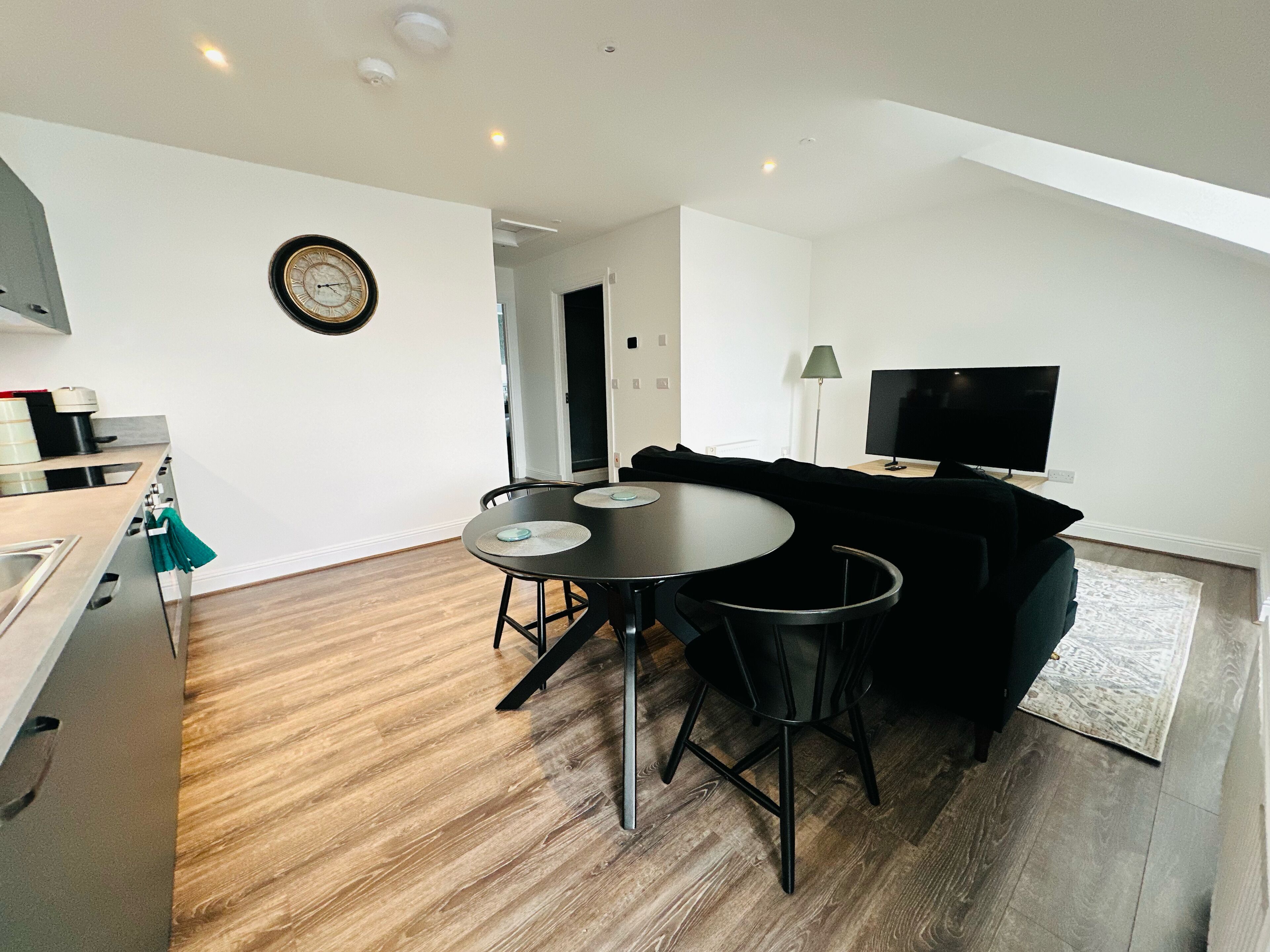 A modern living space with a dining area, a black sofa, a TV, and a kitchen area with a sink and stove.
