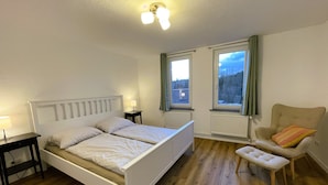 Quarto com cama, cadeira, mesa e duas janelas com cortinas.