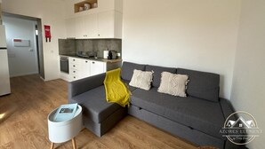 A modern living room with a grey sofa, a small white side table, and a kitchen area in the background. - Azores Element - House with pool (Ponta Delgada)