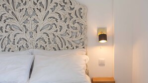 A neatly made bed with white linens, a decorative headboard, a bedside table with a lamp, and a wall-mounted light fixture. - Azores Element - House with pool (Ponta Delgada)