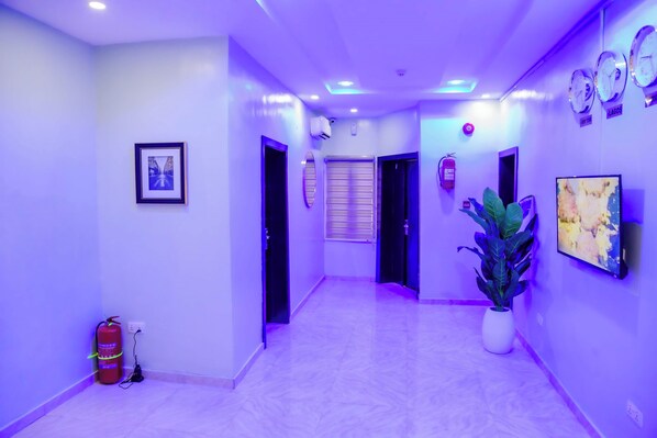 A hallway with a fire extinguisher, a framed picture, a potted plant, and a wall-mounted clock.