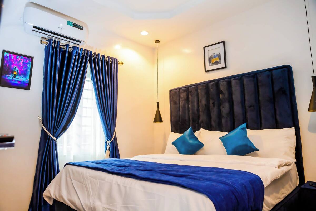 Habitación de hotel con cama, cortinas azules, aire acondicionado montado en la pared, un cuadro enmarco y una lámpara de noche.