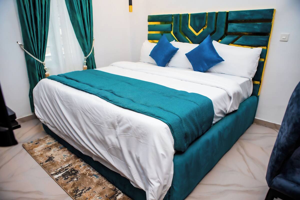 Una cama bien hecha con un edredón verde azulado y blanco, almohadas azules y un cabecero verde.