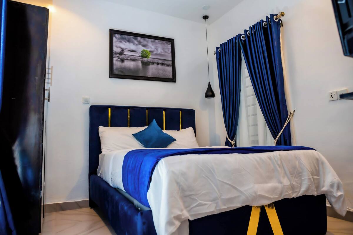 Un dormitorio con una cama, cabecera azul, colcha blanca y azul, almohada azul, un cuadro enmarcado en la pared y cortinas azules.