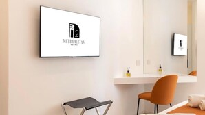 A modern hotel room with a large mirror, a flat-screen TV mounted on the wall, a small table, and an orange chair. - Metropolitan Rooms (Cagliari)