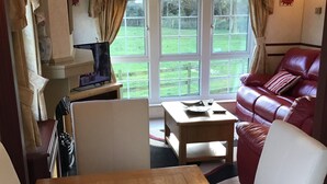 A cozy living area with a wooden table, white chairs, a red leather sofa, and a view of a green landscape through large windows. - Beautiful 2-bed Chalet in Llanfairpwllgwyngyll (Llanfairpwllgwyngyll)
