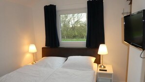 Ein Schlafzimmer mit einem Bett, zwei Nachttischlampen, einem an der Wand befestigten Fernseher und einem Fenster mit Vorhängen.