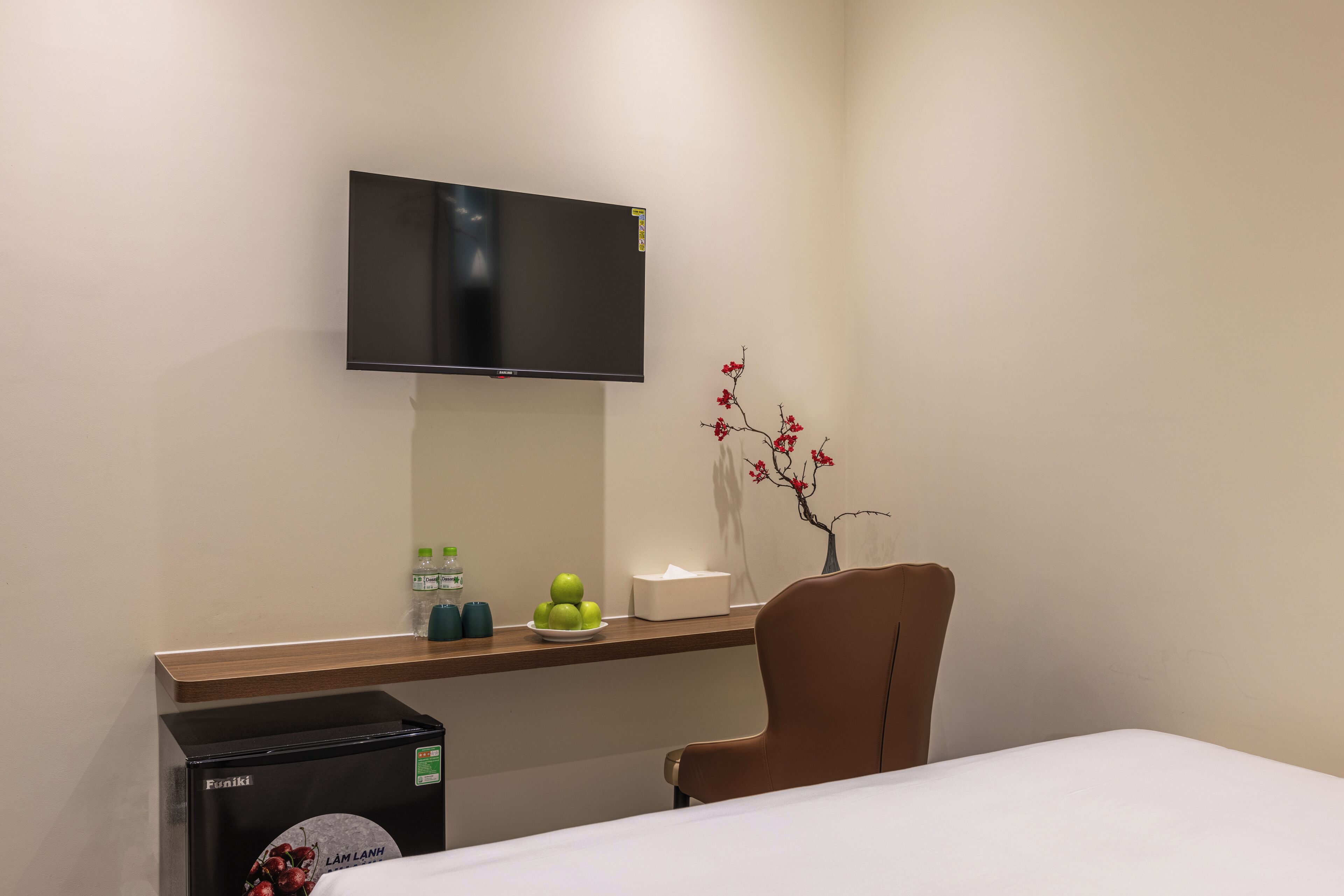 Ein Hotelzimmer mit Bett, Schreibtisch mit Flachbildfernseher, Stuhl, kleinem Kühlschrank, Blumen in einer Vase und einer Wasserflasche.