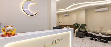 A hotel reception area with a modern design, featuring a white reception desk with a gold script logo, a crescent-shaped light fixture, and a seating area with sofas and a coffee table.