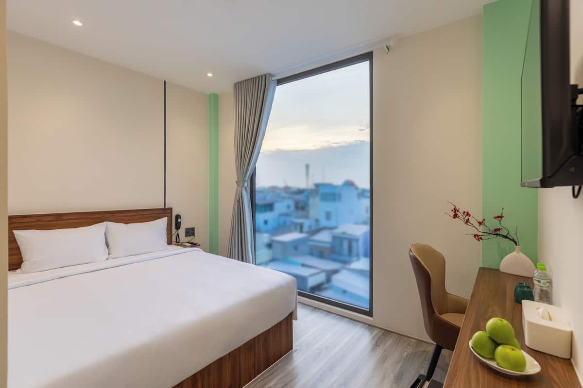 A hotel room with a bed, a desk with a chair, a window with a view of buildings, and a vase with flowers.