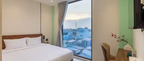 A hotel room with a bed, a desk with a chair, a window with a view of buildings, and a vase with flowers.
