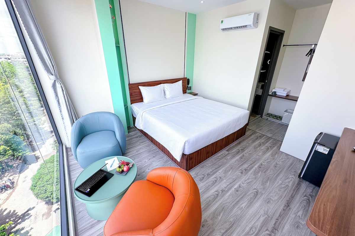 A hotel room with a bed, a desk, a chair, a small table with a laptop, and a view of greenery outside.